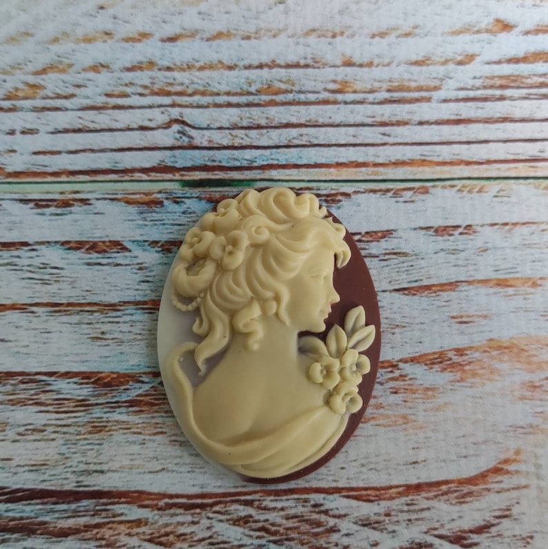 May include: A white cameo with a brown background featuring a woman's profile with curly hair and a floral design.