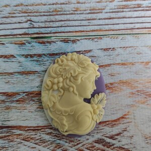 May include: A white cameo with a detailed carving of a woman's profile with a floral design. The cameo is set in a purple oval frame.