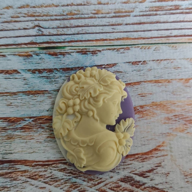 May include: A white cameo with a detailed carving of a woman's profile with a floral design. The cameo is set in a purple oval frame.