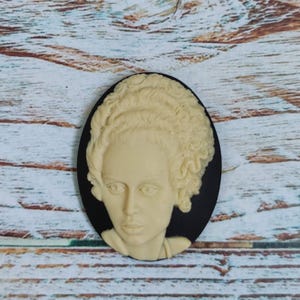 Op de afbeelding: Een zwarte cameo met een wit gesculpteerd beeld van een vrouw met krullend haar. De cameo is ovaalvormig en heeft een zwarte achtergrond.