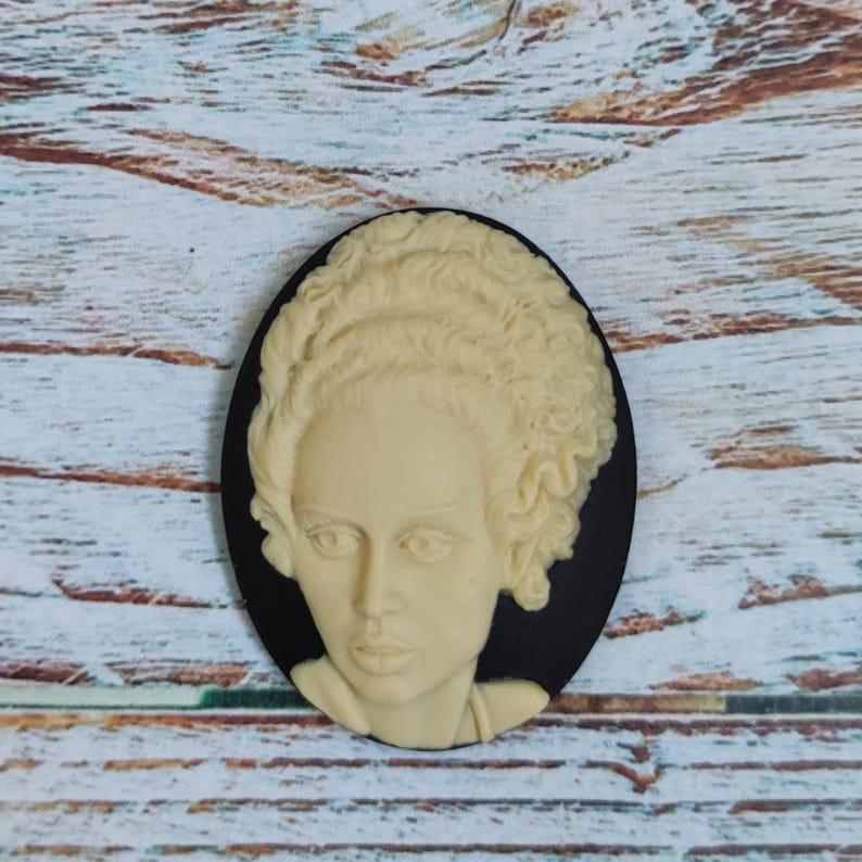 Op de afbeelding: Een zwarte cameo met een wit gesculpteerd beeld van een vrouw met krullend haar. De cameo is ovaalvormig en heeft een zwarte achtergrond.
