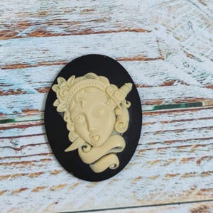 Op de afbeelding: Een zwarte cameo met een witte cameo van een vrouwenhoofd met een dolk door haar hoofd en een banner onder haar kin.