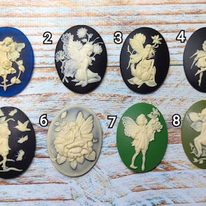 May include: Eight oval-shaped cameo pendants featuring white fairy figurines with wings, each set against a different colored background: blue, black, green, and black. The fairies are depicted in various poses, some with flowers, some with butterflies, and some with other floral details.