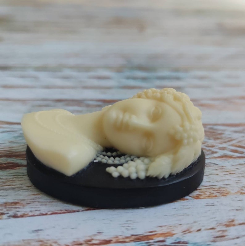 May include: A white cameo featuring a woman's face with a beaded crown, set on a black circular base.