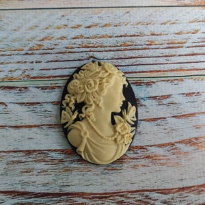 May include: A cream-colored cameo featuring a woman's profile with detailed hair and floral accents. The cameo is set against a black background.