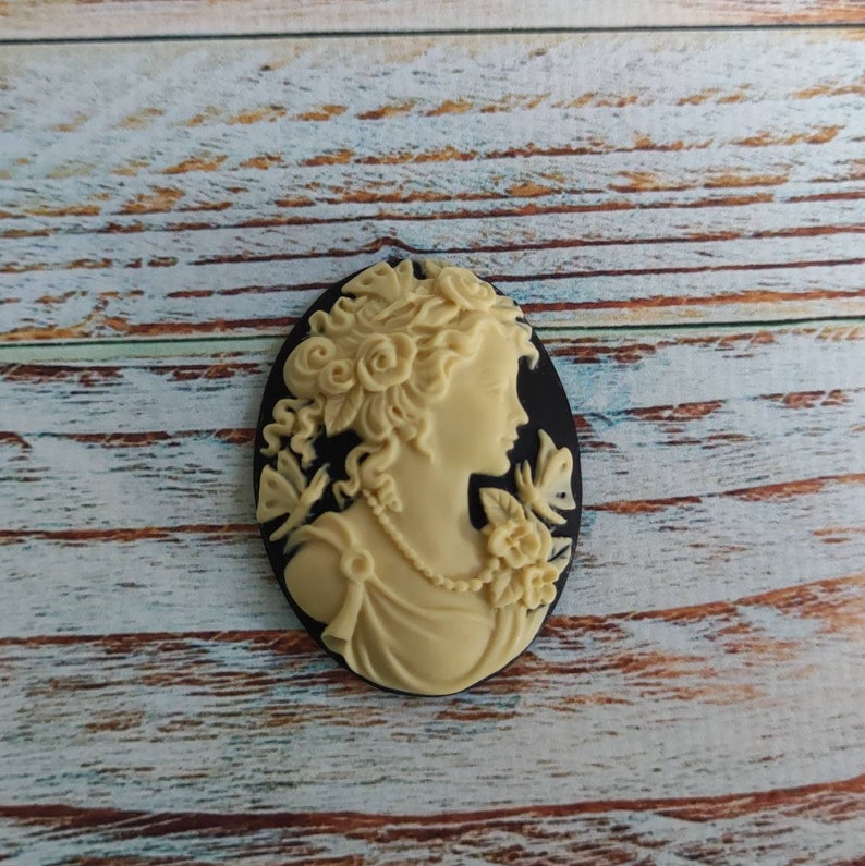 May include: A cream-colored cameo featuring a woman's profile with detailed hair and floral accents. The cameo is set against a black background.
