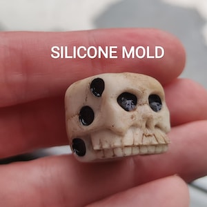 May include: A small, off-white skull-shaped silicone mold with black eye and dot details. The text "SILICONE MOLD" is printed in white above the skull. The mold is held in a person's hand.