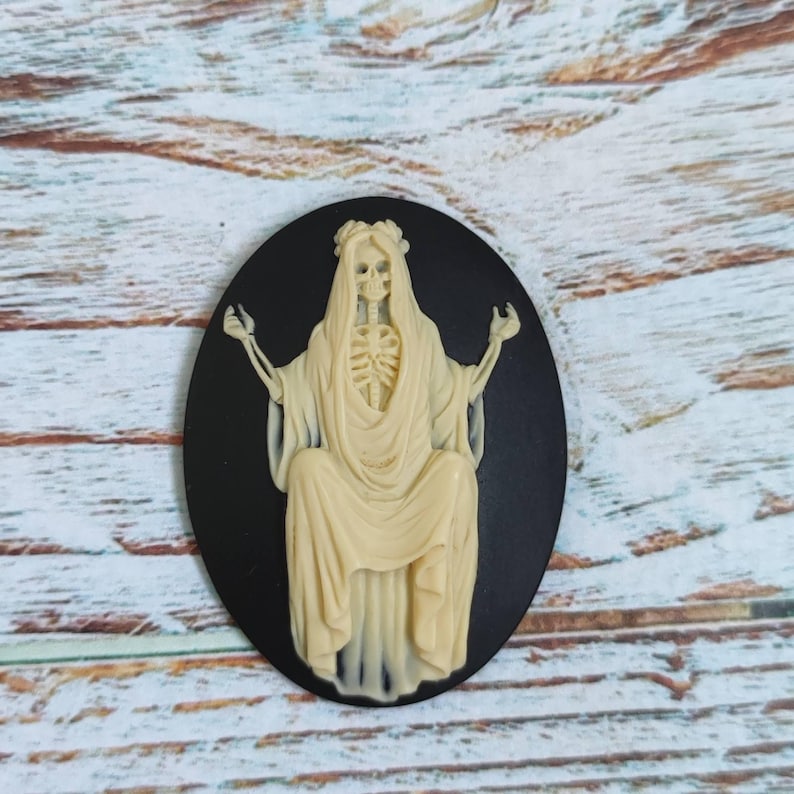 Op de afbeelding: Een zwarte cameo met een wit skeletfiguur dat een gewaad en een bloemenkrans draagt. De figuur zit op een troon met zijn armen uitgestrekt.