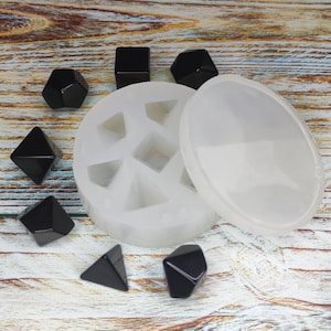 May include: A clear, flexible silicone mold with geometric shapes, including triangles, squares, and pentagons. Several small, black, faceted resin pieces are scattered around the mold. The mold is open, with the lid next to it, ready for resin casting.