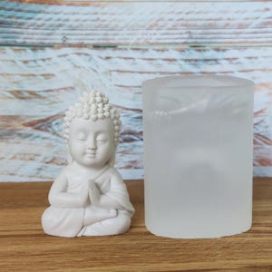 Op de afbeelding: Een witte Boeddha-beeldje met handen in gebedshouding, naast een matglazen kandelaar. De Boeddha heeft een serene uitdrukking en gedetailleerd haar. De achtergrond is een houten oppervlak.