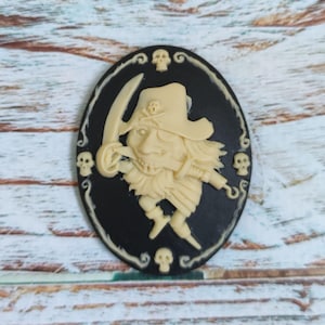 Op de afbeelding: Een zwarte cameo met een wit piraten-schedel en kruisbeenderen ontwerp. De piraat draagt ​​een hoed en heeft een haak voor een hand. De cameo is omgeven door een zwarte rand met witte schedels en krullen.