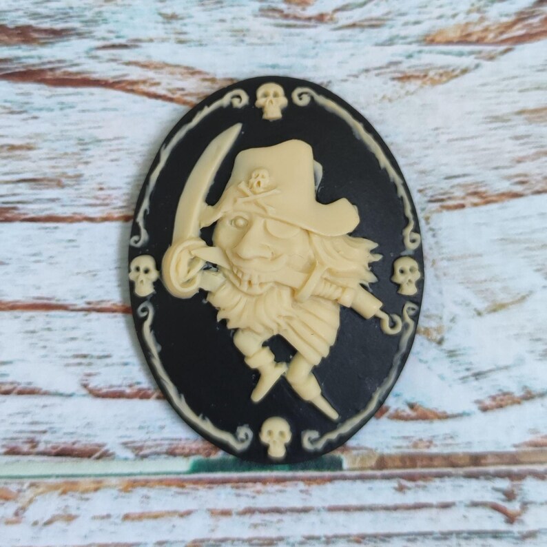 Op de afbeelding: Een zwarte cameo met een wit piraten-schedel en kruisbeenderen ontwerp. De piraat draagt ​​een hoed en heeft een haak voor een hand. De cameo is omgeven door een zwarte rand met witte schedels en krullen.