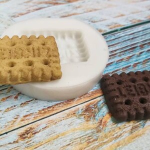 May include: A silicone mold for making small, rectangular cookies. The mold is white and has a raised design of a cookie with a textured surface. Two cookies are shown next to the mold, one is a light brown cookie with a textured surface and the other is a dark brown cookie with a textured surface.