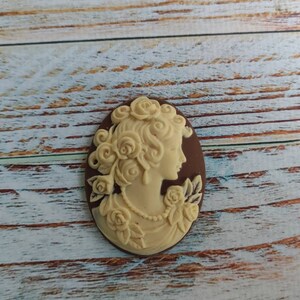 May include: A white cameo with a detailed profile of a woman with a floral design. The cameo is set in a brown oval frame.