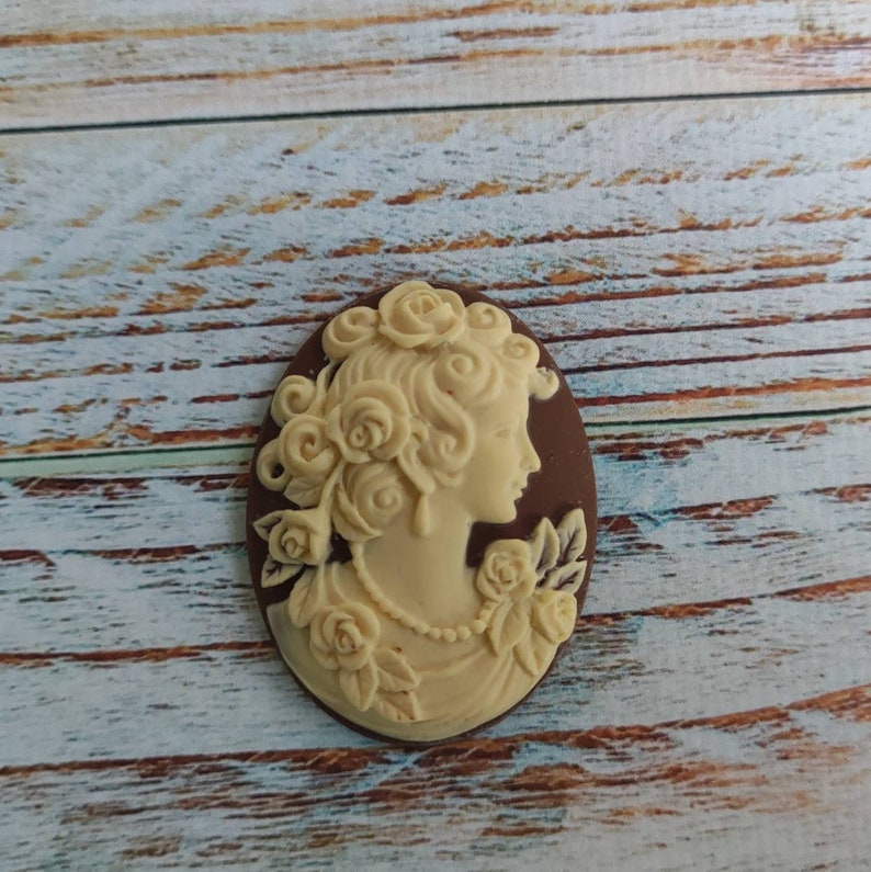 May include: A white cameo with a detailed profile of a woman with a floral design. The cameo is set in a brown oval frame.
