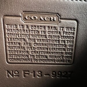 以下が含まれることがあります： 「This is a Coach bag. It was handcrafted in China from completely natural glove-tanned leather. The variations in the grain are characteristic of natural full-grain leather. Its superior craftsmanship and attention to detail reflect our commitment to enduring quality.」というテキストと「№ F13-9927」という番号が書かれた、茶色の革製のコーチバッグのタグのクローズアップ。
