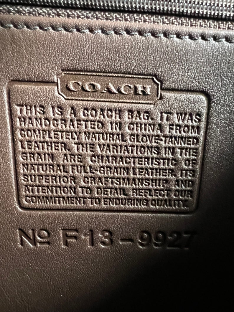以下が含まれることがあります： 「This is a Coach bag. It was handcrafted in China from completely natural glove-tanned leather. The variations in the grain are characteristic of natural full-grain leather. Its superior craftsmanship and attention to detail reflect our commitment to enduring quality.」というテキストと「№ F13-9927」という番号が書かれた、茶色の革製のコーチバッグのタグのクローズアップ。