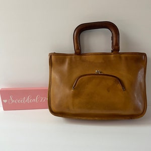 May include: A brown leather tote bag with a wooden handle. The bag has a flap closure with a gold clasp.