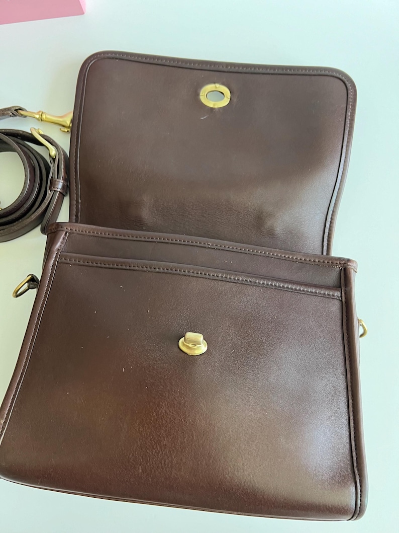 May include: Open, brown leather shoulder bag with a flap closure and gold-tone hardware. The bag has a long, adjustable strap and a spacious interior. The bag's design is classic and timeless, suitable for everyday use.