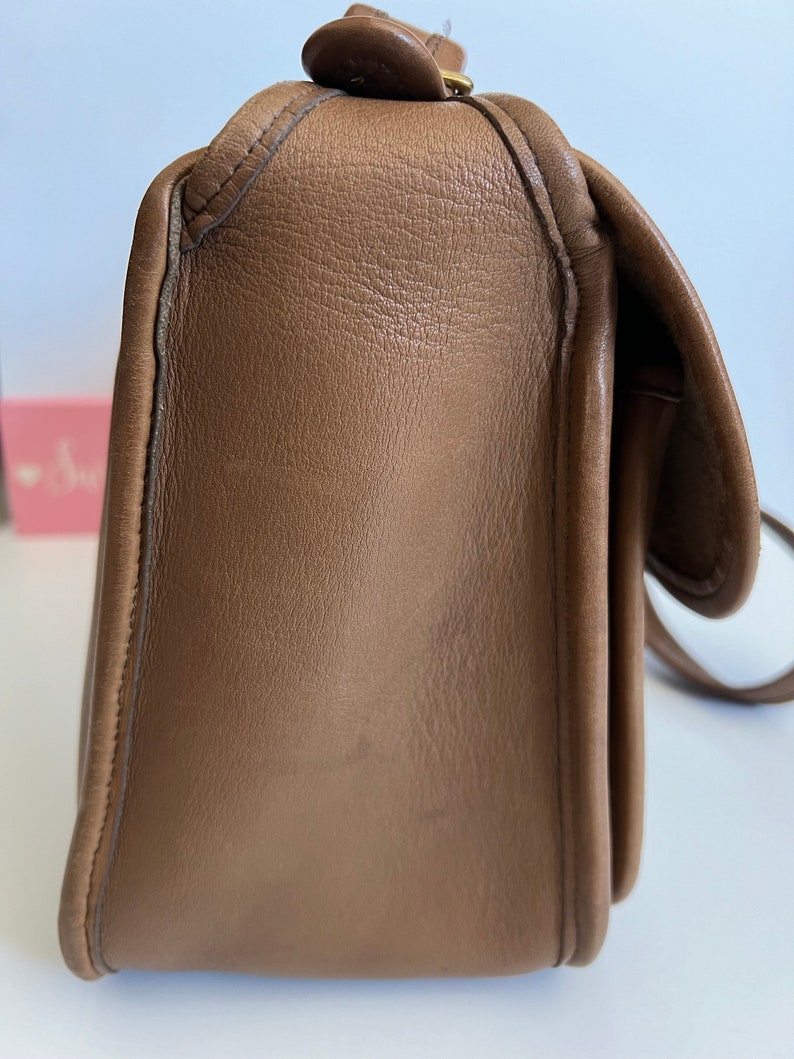 May include: A brown leather shoulder bag with a flap closure and a gold-toned buckle. The bag has a smooth, textured surface and a slightly rounded shape.