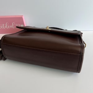 May include: A dark brown leather crossbody bag with a flap closure and a gold-tone clasp. The bag has a rectangular shape and a shoulder strap. The bag is a vintage style.