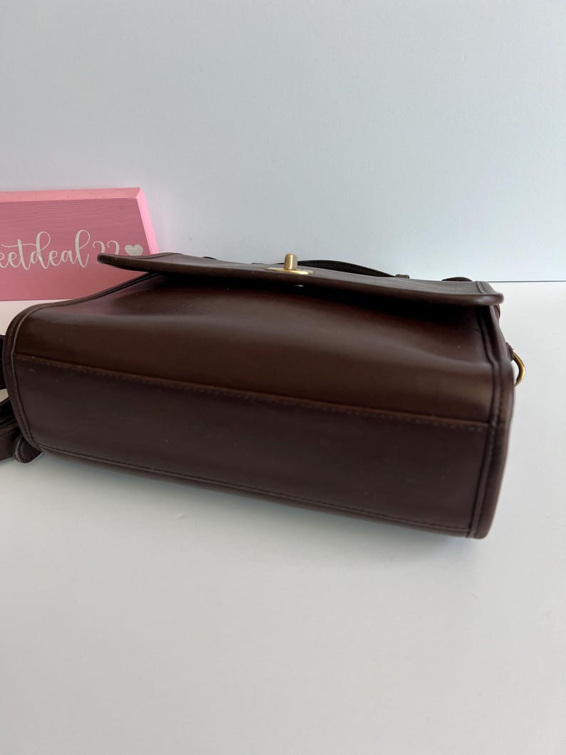 May include: A dark brown leather crossbody bag with a flap closure and a gold-tone clasp. The bag has a rectangular shape and a shoulder strap. The bag is a vintage style.