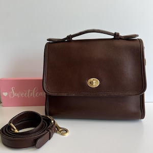 May include: A dark brown leather satchel with a top handle and a detachable shoulder strap. The bag features a gold-tone clasp and hardware. The strap is coiled next to the bag. A pink box with the text "Sweetdea" is in the background.