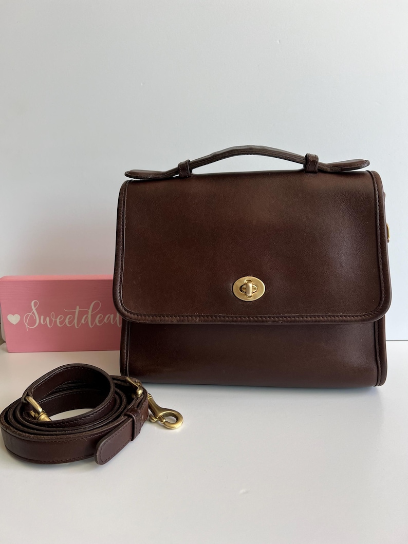 May include: A dark brown leather satchel with a top handle and a detachable shoulder strap. The bag features a gold-tone clasp and hardware. The strap is coiled next to the bag. A pink box with the text "Sweetdea" is in the background.