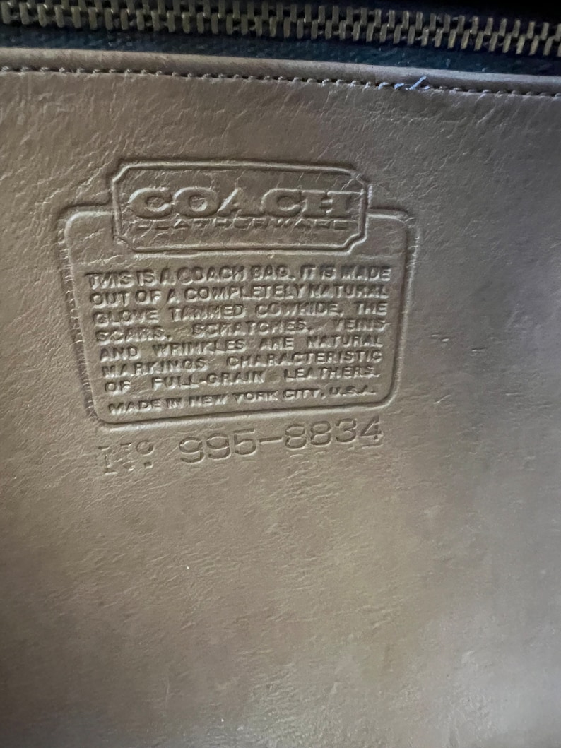 May include: Close-up of a brown leather Coach bag tag with the text "This is a Coach bag. It is made out of a completely natural glove tanned cowhide. The scars, scratches, veins and wrinkles are natural markings characteristic of full-grain leathers. Made in New York City, USA." and the number "1995-8834".