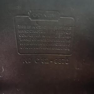 May include: Close-up of a black leather Coach label. The label reads "THIS IS A COACH... HAND CRAFTED... COMPLETELY NATURAL GLOVE... IN THE GRAIN... NO C82-9870."