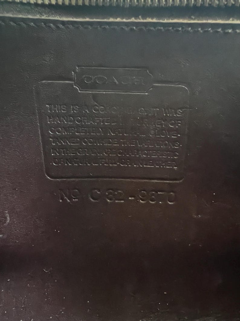 May include: Close-up of a black leather Coach label. The label reads "THIS IS A COACH... HAND CRAFTED... COMPLETELY NATURAL GLOVE... IN THE GRAIN... NO C82-9870."