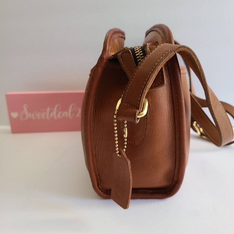 May include: A brown leather crossbody bag with a gold zipper and a gold strap. The bag has a small, rectangular tag with the word "Coach" on it.