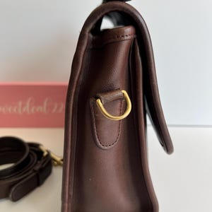 May include: A dark brown leather handbag with a flap closure. The bag features a gold-colored metal ring and a matching strap. The leather appears to be smooth and well-maintained, with a classic design.