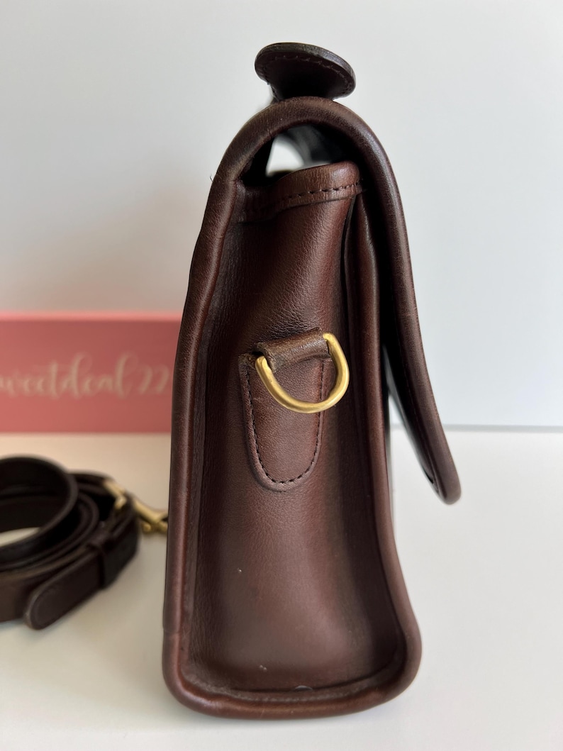 May include: A dark brown leather handbag with a flap closure. The bag features a gold-colored metal ring and a matching strap. The leather appears to be smooth and well-maintained, with a classic design.