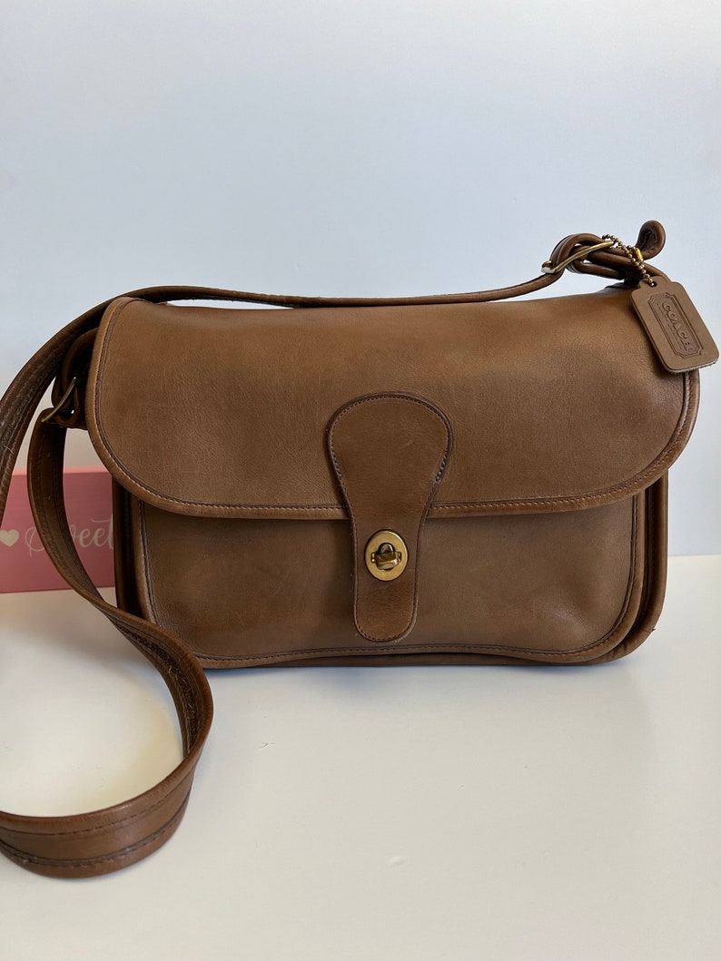 May include: A brown leather shoulder bag with a flap closure and a gold-toned buckle. The bag has a long adjustable strap and a small leather tag with the word "COACH" embossed on it.