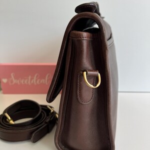 May include: A dark brown leather satchel with a flap closure and a detachable shoulder strap. The bag features gold-toned hardware, including a D-ring for the strap. The strap is coiled next to the bag. The bag is on a white surface.
