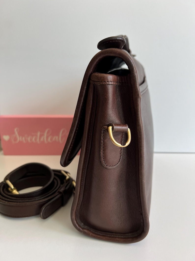 May include: A dark brown leather satchel with a flap closure and a detachable shoulder strap. The bag features gold-toned hardware, including a D-ring for the strap. The strap is coiled next to the bag. The bag is on a white surface.