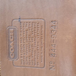 May include: Close-up of a brown leather Coach bag tag with the text "This is a Coach bag. It is made out of a completely natural glove tanned cowhide, the scars, scratches, veins and wrinkles are natural markings characteristic of full-grain leathers. Made in the United States" and the number "N&deg; 444-9344".
