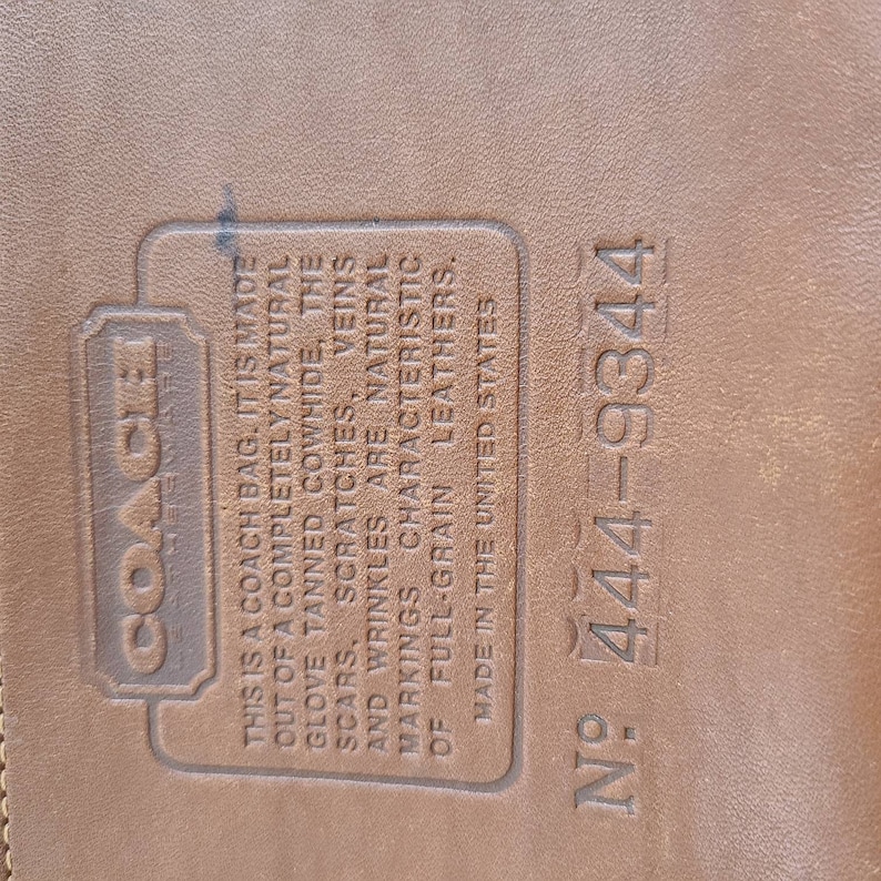May include: Close-up of a brown leather Coach bag tag with the text "This is a Coach bag. It is made out of a completely natural glove tanned cowhide, the scars, scratches, veins and wrinkles are natural markings characteristic of full-grain leathers. Made in the United States" and the number "N&deg; 444-9344".