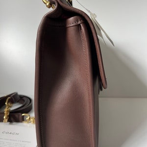 以下が含まれることがあります： 金色の留め具とストラップが付いた、茶色の革製のショルダーバッグ。バッグはフラップで閉じられ、前面に小さなポケットがあります。バッグはコーチ製です。