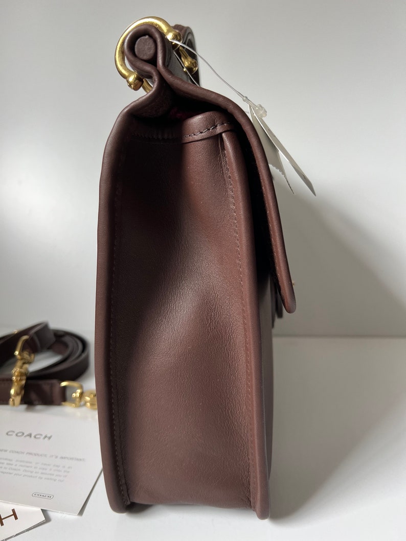 以下が含まれることがあります： 金色の留め具とストラップが付いた、茶色の革製のショルダーバッグ。バッグはフラップで閉じられ、前面に小さなポケットがあります。バッグはコーチ製です。