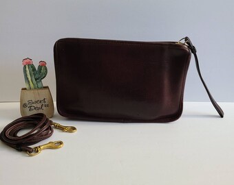 May include: A dark brown leather rectangular clutch bag with a zipper closure and a wrist strap. A matching detachable leather shoulder strap with brass swivel clips is included. The bag features a smooth finish and a simple, structured design.
