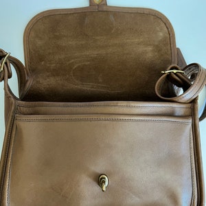 May include: A brown leather shoulder bag with a flap closure and a gold-toned buckle. The bag has a suede lining and a single pocket on the front.