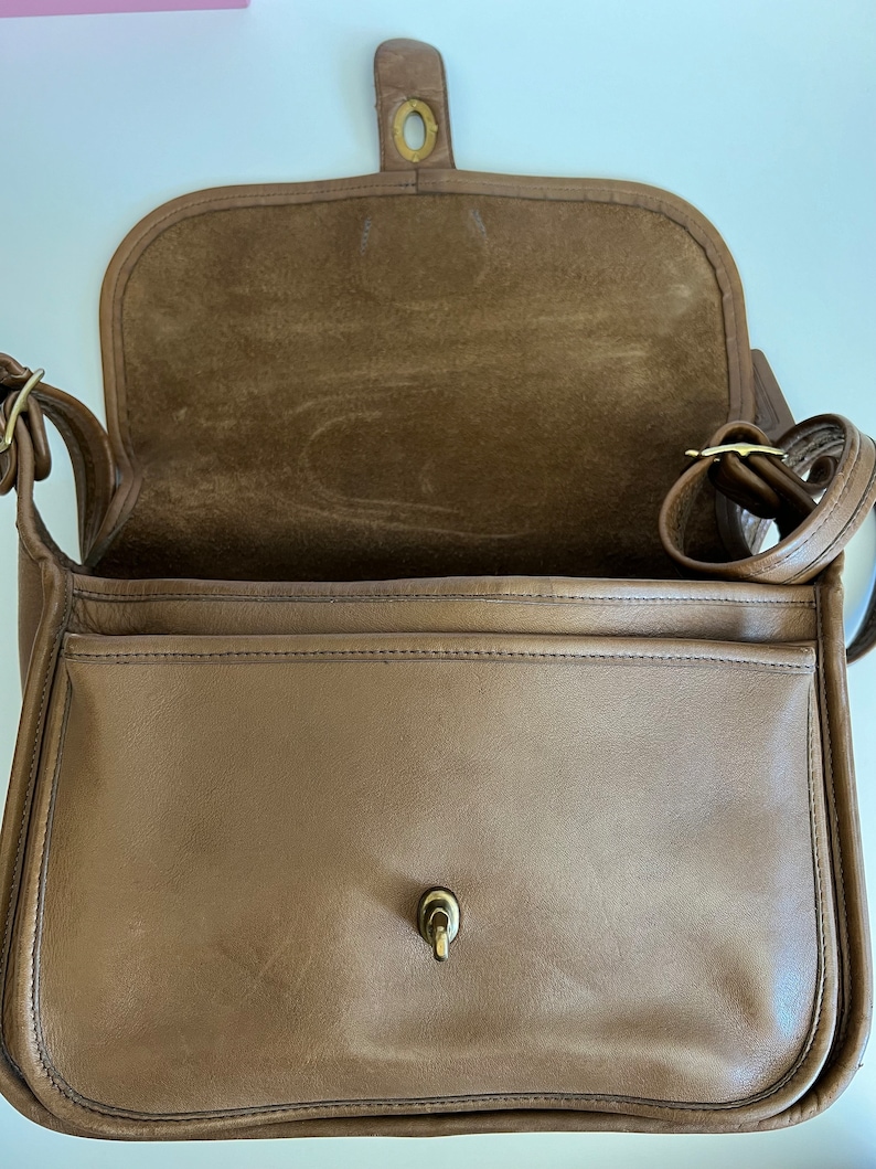 May include: A brown leather shoulder bag with a flap closure and a gold-toned buckle. The bag has a suede lining and a single pocket on the front.