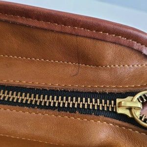 May include: Close-up of a brown leather bag with a black zipper and gold zipper pull.