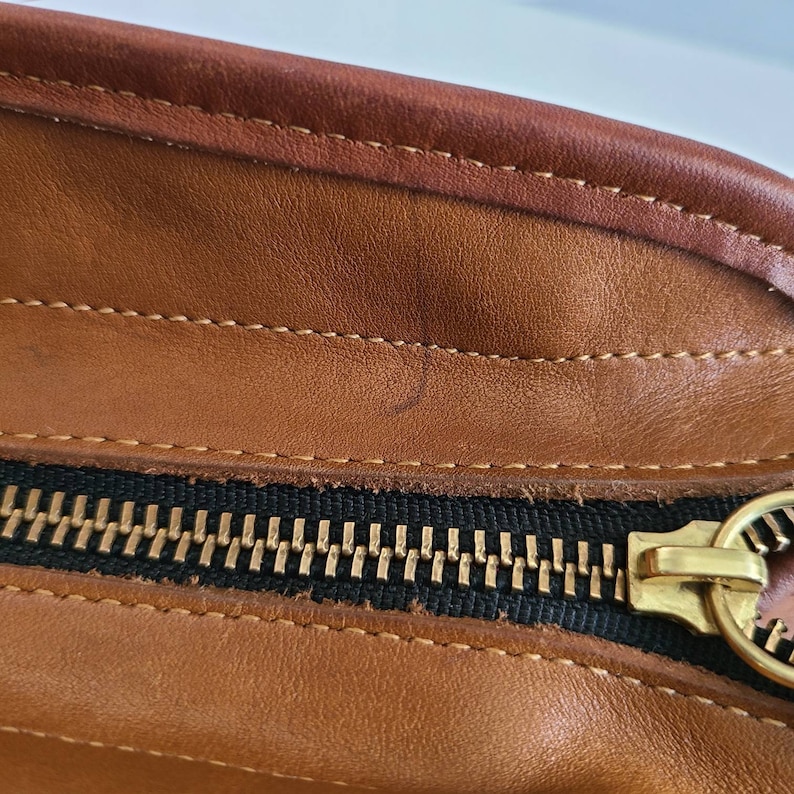 May include: Close-up of a brown leather bag with a black zipper and gold zipper pull.