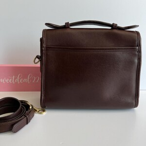 May include: A dark brown leather satchel bag with a top handle and a detachable shoulder strap. The bag has a rectangular shape with a front pocket and gold-tone hardware. The words "sweetdeal22" are visible on a pink box in the background.