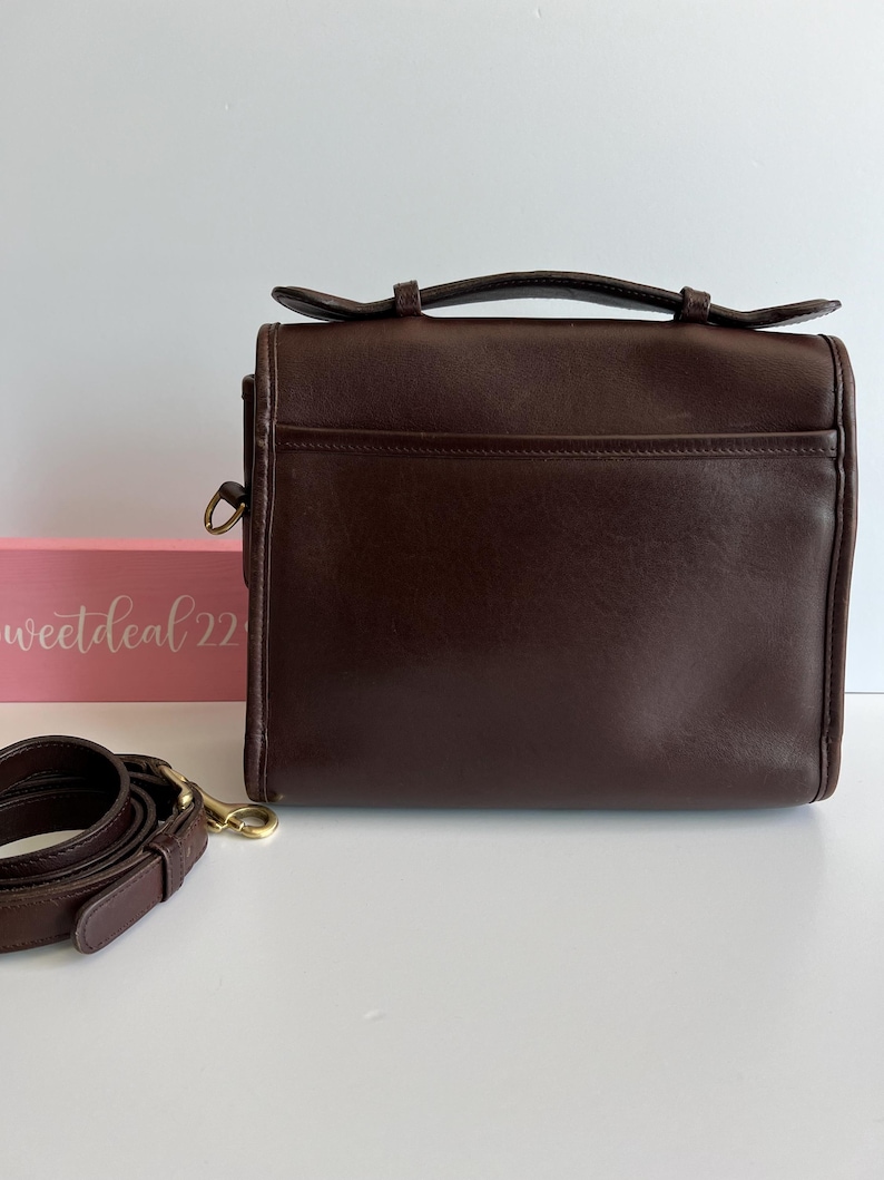May include: A dark brown leather satchel bag with a top handle and a detachable shoulder strap. The bag has a rectangular shape with a front pocket and gold-tone hardware. The words "sweetdeal22" are visible on a pink box in the background.