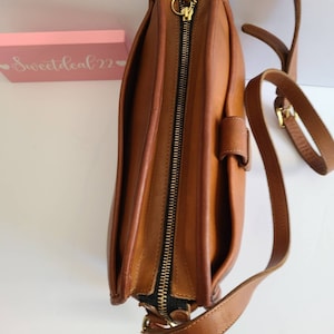 May include: A brown leather satchel bag with a zipper closure and a long adjustable strap. The bag has a flap with a buckle closure.