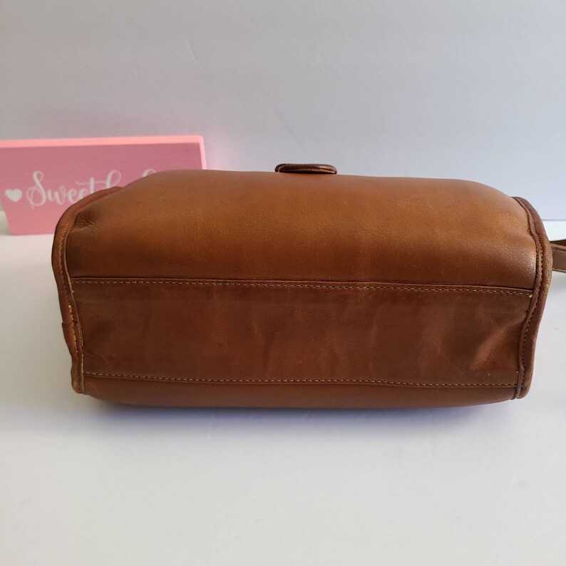 May include: A brown leather handbag with a single buckle closure. The bag is shown from the bottom, with the stitching visible.